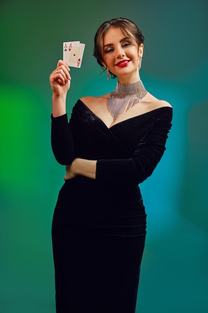 Fille brune élégante, épaules nues et maquillage lumineux. En robe noire, collier scintillant et boucles d'oreilles. Souriant, montrant deux cartes à jouer, posant sur fond coloré. Poker, casino. Fermer