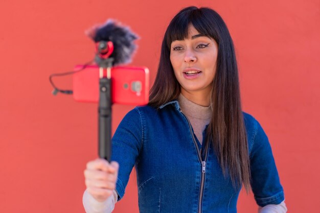 Fille brune enregistrant un vlog dans une tenue en denim bleu. Jolie caucasienne un fond rouge