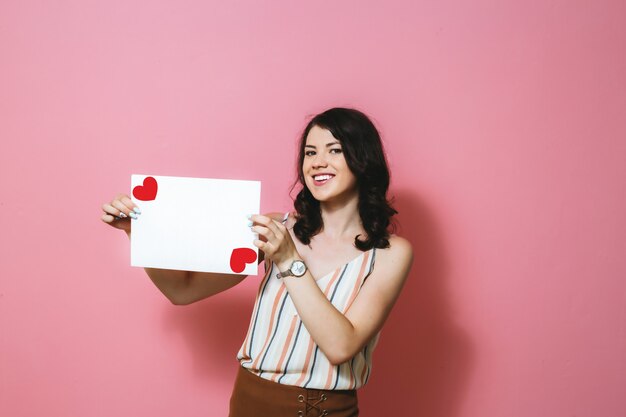 Une fille brune heureuse tenant un blanc blanc avec un coeur sur un mur rose