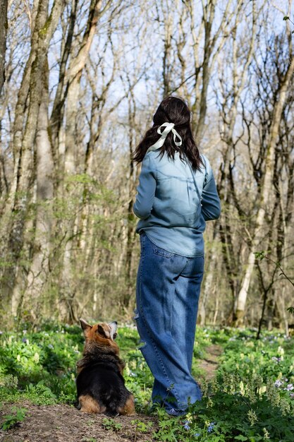 une fille brune en jeans et une chemise en jean lors d'une promenade dans la forêt avec un chien corgi