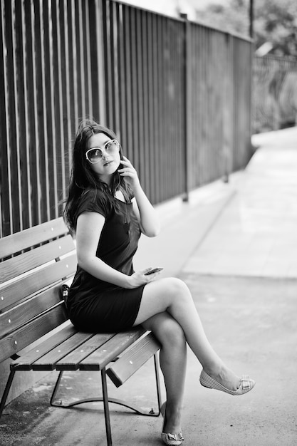 Fille brune à lunettes de soleil robe noire assis sur un banc en écoutant de la musique à partir d'un téléphone casque et posant dans la rue de la ville