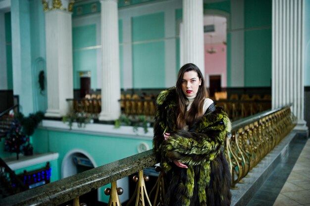 Fille brune en manteau de fourrure vert dans le vieux hall avec colonne et garde-corps