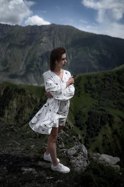 Une fille brune mince en blanc pose et regarde au loin sur fond de montagnes