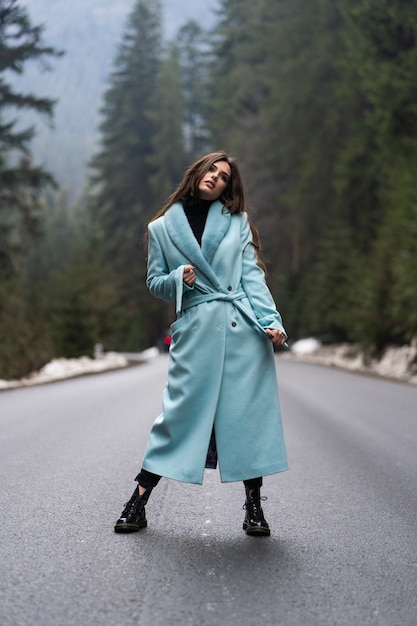 Fille brune sexy poasing sur route sur la table des montagnes d'hiver. Glamour jeune femme portant un long manteau bleu élégant. Concept de mode, d'affaires et de belles personnes.