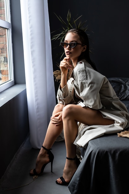 Fille brune sexy portant un trench-coat et de la lingerie sur le lit