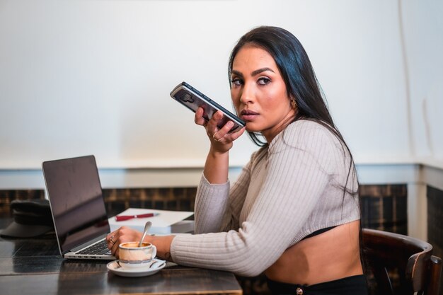 Fille brune télétravaillant dans une cafétéria avec son ordinateur portable, envoyant un message audio à un collègue