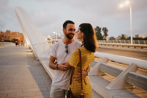 Une fille brune vêtue d'une robe jaune et son petit ami souriant s'embrassent sur un pont blanc à Valence. Un couple de touristes à un rendez-vous dans la chaude soirée.
