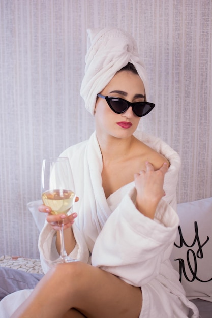 Fille buvant du champagne et lisant un magazine Jolie femme dans la chambre en peignoir blanc se trouve dans son lit en buvant du vin et tourne les pages du magazine de mode Vue rapprochée