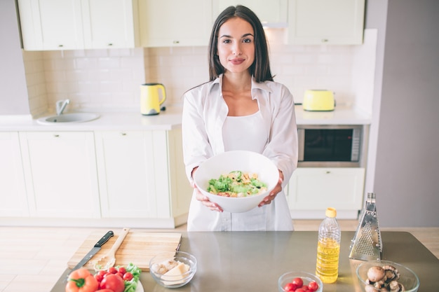 Une fille calme et paisible se lève et montre un bol avec de la salade. Elle a l'air. Il y a des ingrédients sur la table qui ont été laissés de la cuisson.