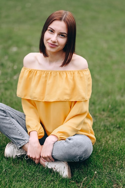 Une fille avec un carré et un chemisier jaune est assise sur l'herbe verte et sourit photo d'en haut