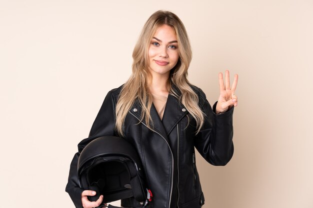 fille avec un casque de moto sur le mur beige heureux et en comptant trois avec les doigts