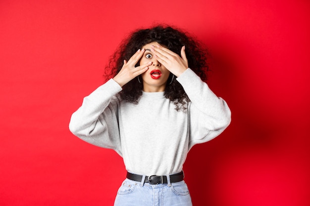 Fille caucasienne choquée couvrant les yeux et regardant à travers les doigts quelque chose d'incroyable, haletant sans voix, debout sur fond rouge