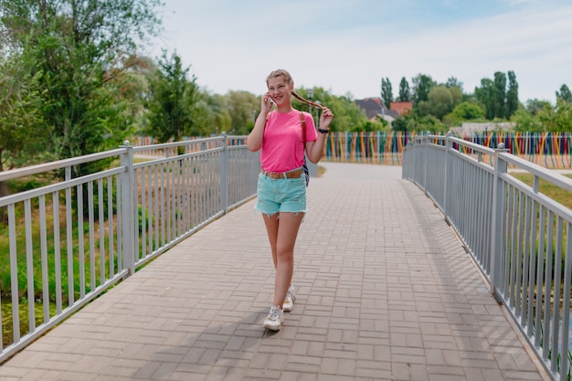 Fille caucasienne dans un T-shirt rose parlant au téléphone. Une étudiante souriante sourit sur la route.