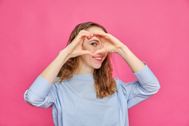 Fille caucasienne élégante dans un t-shirt bleu pâle sur fond rose. Les mains montrent le cœur.