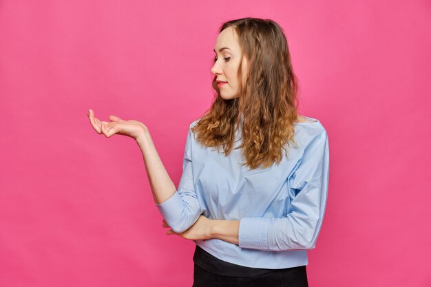 Fille caucasienne élégante dans un t-shirt bleu pâle en regardant un objet abstrait dans la paume de sa main sur un fond rose.