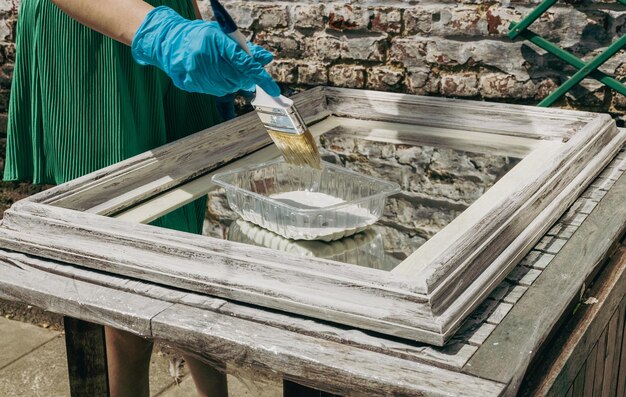 Une fille caucasienne et des gants peignent un cadre de miroir en plongeant un pinceau dans une boîte avec de la peinture blanche