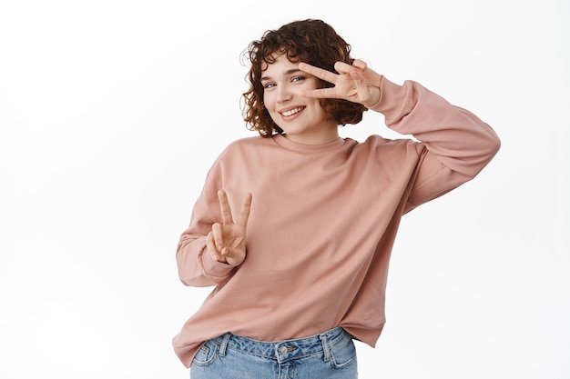 Fille caucasienne heureuse et positive, dansant avec des signes de pièce kawaii, l'air gaie, debout dans des vêtements décontractés sur blanc