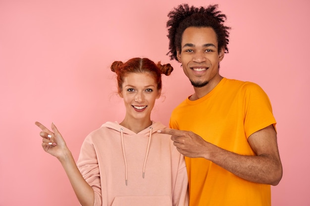 Une fille caucasienne et un mec afro-américain posant sur fond de studio rose pointent le doigt vers le côté gauche