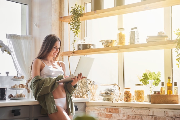 Fille charmante. Une brune heureuse s'appuyant sur ses meubles tout en travaillant sur son ordinateur portable dans la cuisine