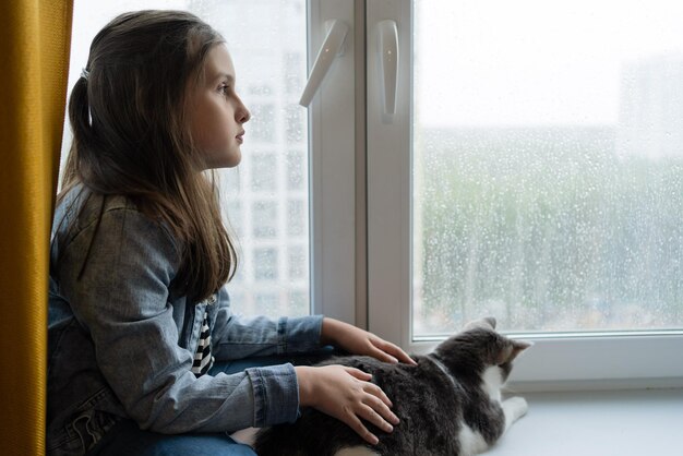 Fille avec un chat regarde par la fenêtre par temps de pluie