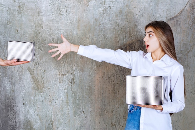 Une fille en chemise blanche debout sur un mur de béton se voit offrir une boîte-cadeau en argent et des mains impatientes de la prendre