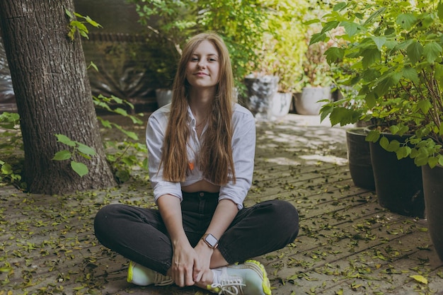 Une fille en chemise blanche est assise près des arbres et des feuilles et regarde la caméra