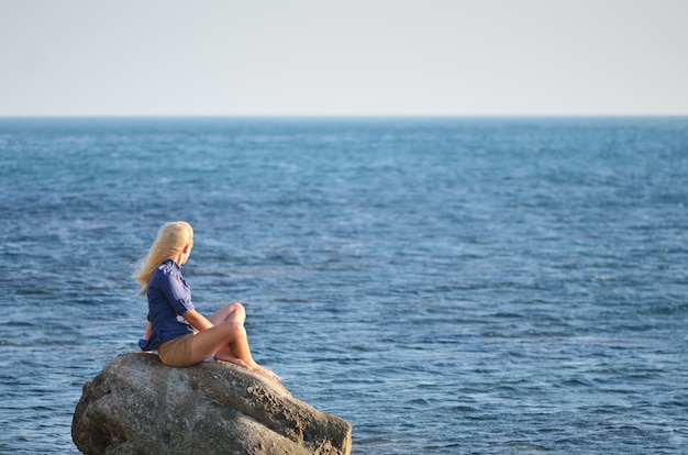 Fille à la chemise bleue assise sur un rocher et regardant la mer