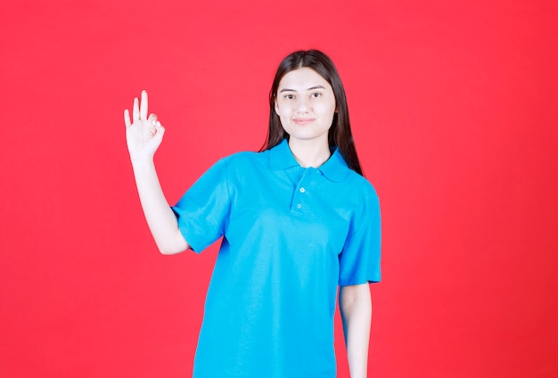 Fille en chemise bleue debout sur un mur rouge et montrant un signe positif de la main