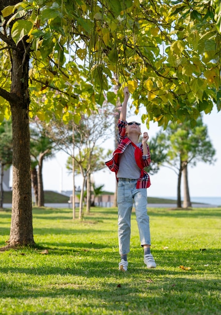 Une fille en chemise à carreaux saute pour les feuilles sur un arbre dans un parc de la ville un jour d'été ensoleillé Bon concept de vacances pour adolescents