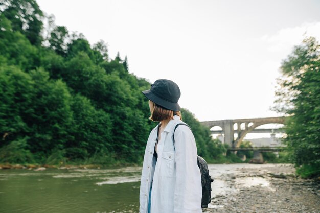 Fille en chemise et chapeau se dresse sur les rives d'une rivière de montagne et sur le fond du pont