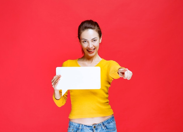 Fille en chemise jaune tenant un panneau d'information rectangle et pointant la personne devant