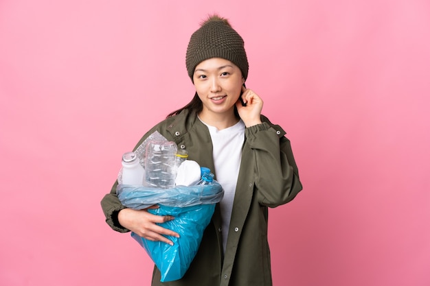 Fille chinoise tenant un sac plein de bouteilles en plastique à recycler sur mur rose isolé en riant