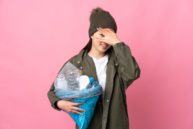 Fille chinoise tenant un sac plein de bouteilles en plastique à recycler sur rose couvrant les yeux par les mains et souriant
