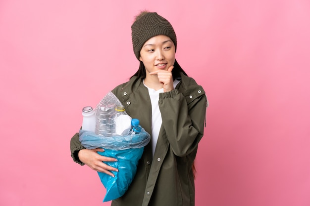 Fille chinoise tenant un sac plein de bouteilles en plastique à recycler sur rose isolé à la recherche sur le côté et souriant