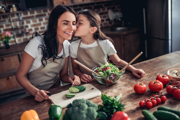 Fille chuchotant sa mère à l'oreille tout en préparant une salade fraîche ensemble dans la cuisine