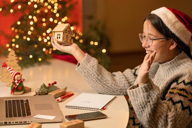 une fille coiffée d'une casquette de Noël est assise devant un ordinateur portable avec une maison en bois dans les mains