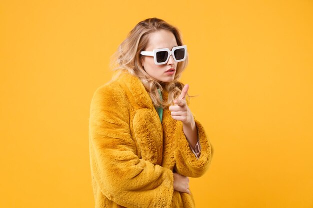 Fille confiante jeune femme blonde en manteau de fourrure jaune, lunettes de soleil foncées posant isolé sur fond orange portrait en studio. Concept de style de vie des gens. Maquette de l'espace de copie. Pointez l'index vers la caméra.