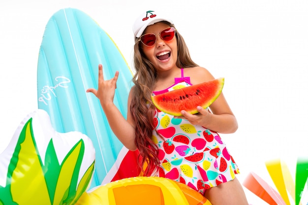 Fille cool avec de longs cheveux crucifiés dans des lunettes de soleil et un maillot de bain se tient près de matelas gonflables avec une pastèque