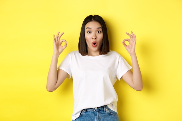 Fille coréenne impressionnée disant WOW, montrant des signes corrects et l'air étonné, debout sur fond jaune