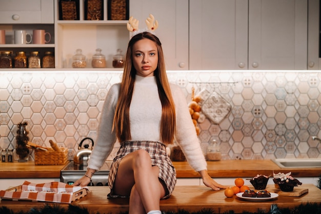 Une fille avec des cornes de renne à Noël est assise sur la table de la cuisine et sourit.Femme au nouvel an dans la cuisine