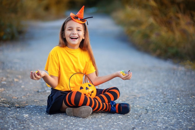 Une fille en costume d'Halloween est assise par terre et savoure les bonbons de son panier. L'enfant se réjouit des bonbons pour Halloween