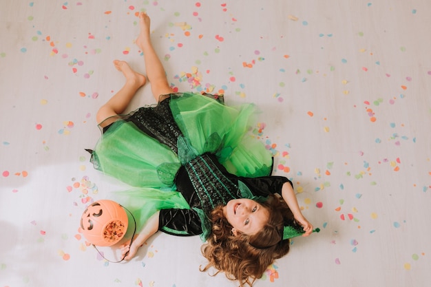 Fille en costume d'Halloween vert d'une sorcière ou d'une fée se trouve sur le sol parsemé de confettis colorés