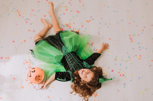 Fille en costume d'Halloween vert d'une sorcière ou d'une fée se trouve sur le sol parsemé de confettis colorés