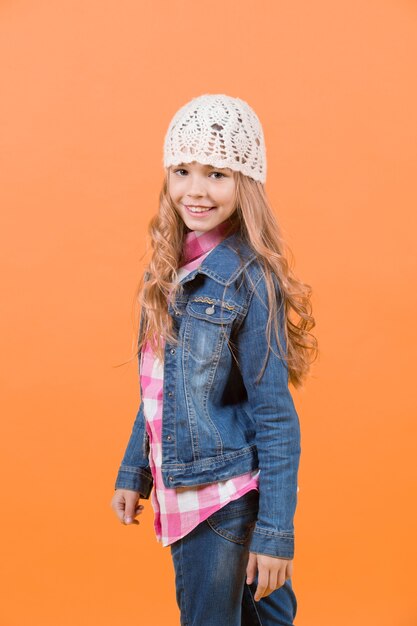 Fille en costume jeans, chapeau, chemise à carreaux sur fond orange. Concept d'enfance heureuse. Sourire d'enfant aux longs cheveux blonds. Beauté, look, coiffure. Mode, style, tendance.