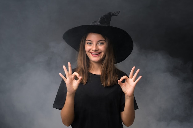 Fille avec costume de sorcière pour les fêtes d'halloween sur un mur noir isolé, montrant un signe ok avec les doigts