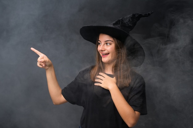 Fille avec costume de sorcière pour les fêtes d'halloween sur un mur noir isolé, pointant le doigt sur le côté