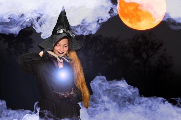 Une fille en costume de sorcière pour halloween évoque une boule lumineuse et brillante Effet magique sur une photo sombre