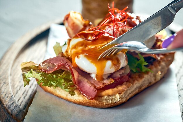 Une fille coupe un sandwich avec du bacon à la tomate et un œuf poché pour le petit-déjeuner Fond de béton Lumière dure