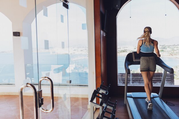 Fille sur la course du matin sur le tapis roulant dans la salle de gym devant une grande fenêtre sur le fond de la mer