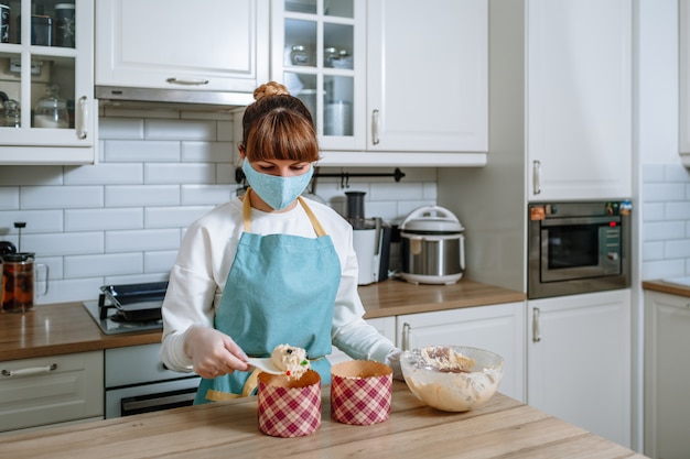 Fille cuisinier dans un masque médical et des gants, étendant la pâte dans des moules pour gâteaux de Pâques